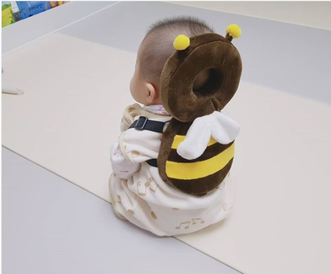 A baby sits on the floor wearing a cute bee-shaped backpack with yellow and black stripes, white wings, and antennae. The baby is dressed in light-colored clothes with a musical note pattern.