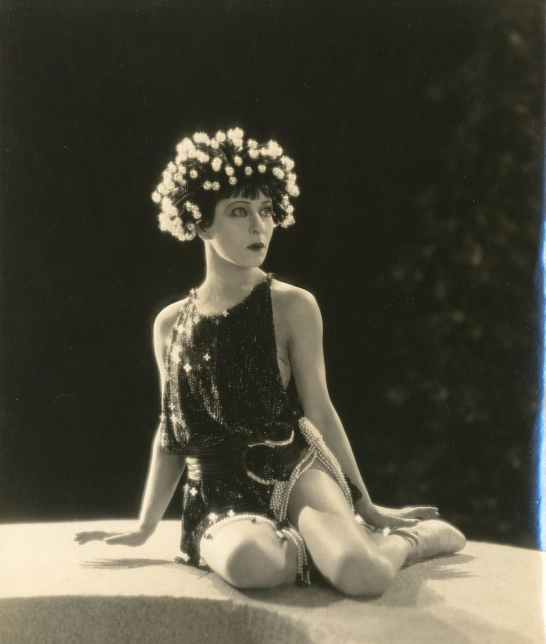 A woman in a sparkling, sleeveless outfit and a headpiece of glowing orbs sits sideways on a round platform, gazing to her left. The photo is in black and white with dramatic lighting.