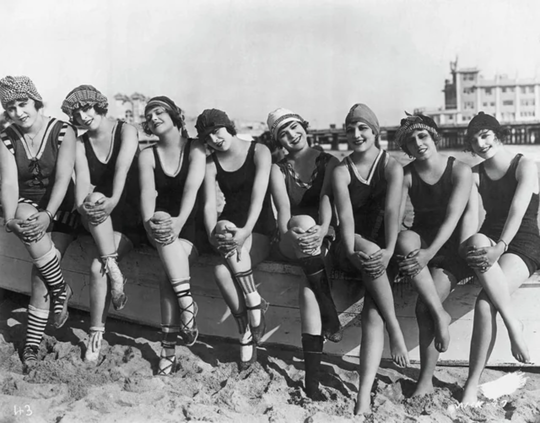 A group of women in vintage swimsuits and swim caps sit on a wooden beam at a sandy beach, smiling and posing together, with a pier and buildings visible in the background.
