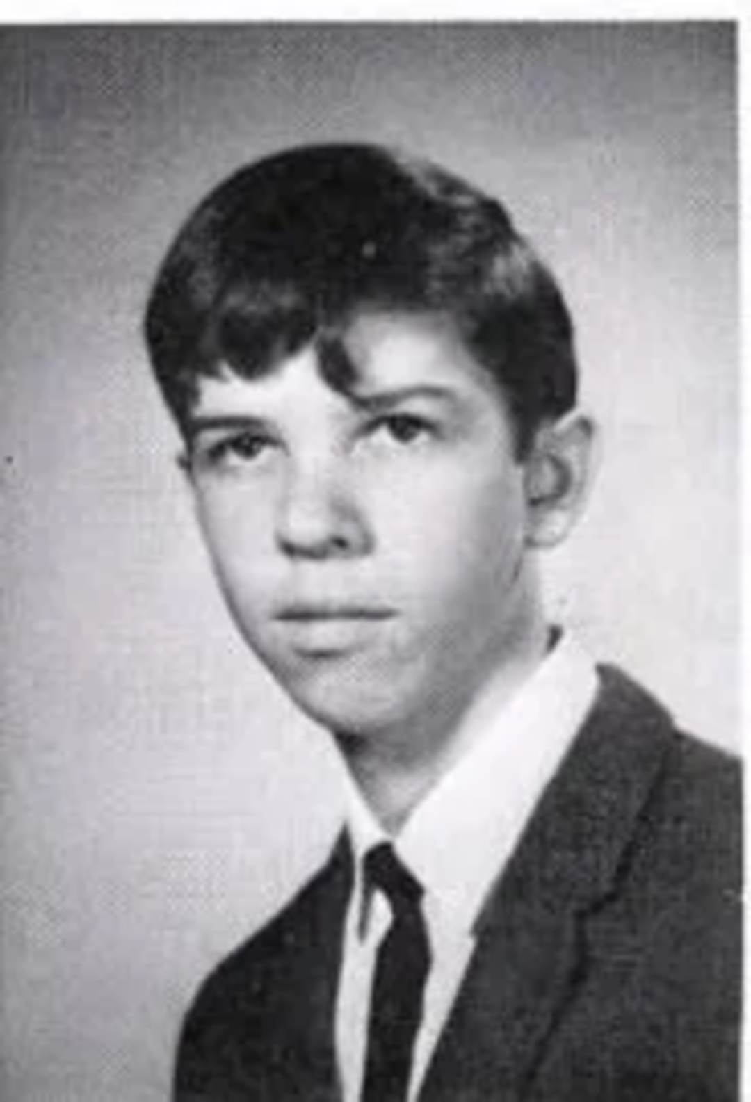 Black and white yearbook-style portrait of a teenage boy with short dark hair, wearing a suit jacket, white shirt, and dark tie, posed against a plain background.