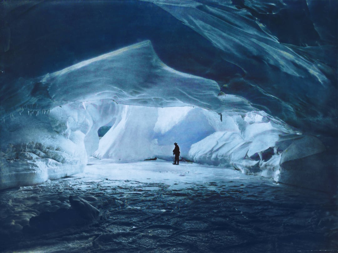 A person stands inside a vast, blue-tinted ice cave with textured walls and ceiling, surrounded by frozen formations and illuminated by natural light filtering through the ice.