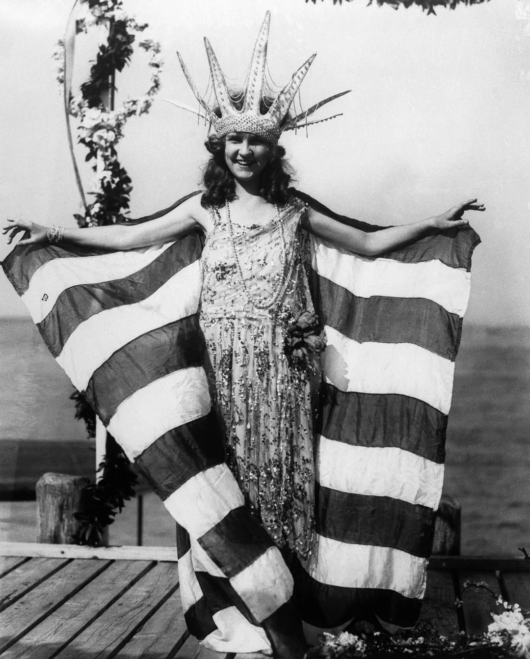 A woman poses outdoors wearing a crown resembling the Statue of Liberty’s and a dress with draped fabric in the pattern of the American flag. She stands on a wooden platform near water, holding the flag open like wings.