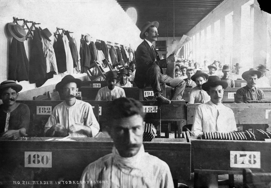 Black-and-white photo of men working at desks in a tobacco factory. A man sits above them reading aloud. Coats hang on wall hooks. Desks are numbered and men look toward the camera, some holding cigars.