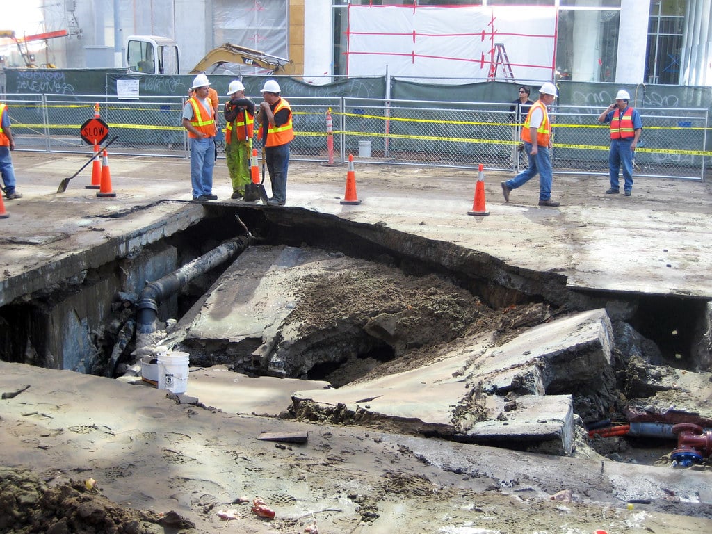 Several construction workers in safety gear stand near a large hole with broken concrete and exposed pipes on a construction site, with safety cones and fencing around the area.