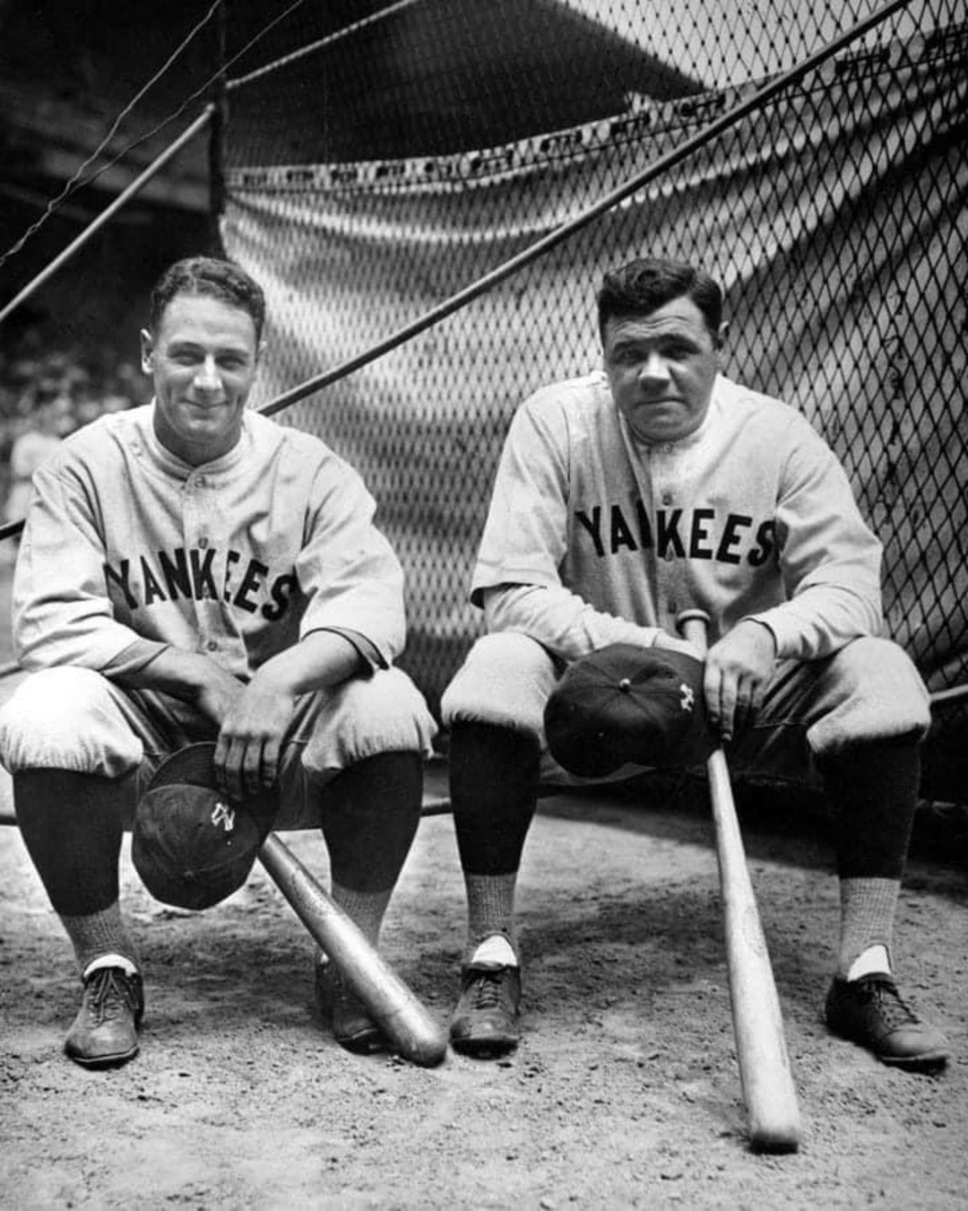Two New York Yankees baseball players sit side by side on a bench in uniform, each holding a bat and resting their caps on their knees, with a netted backdrop behind them.