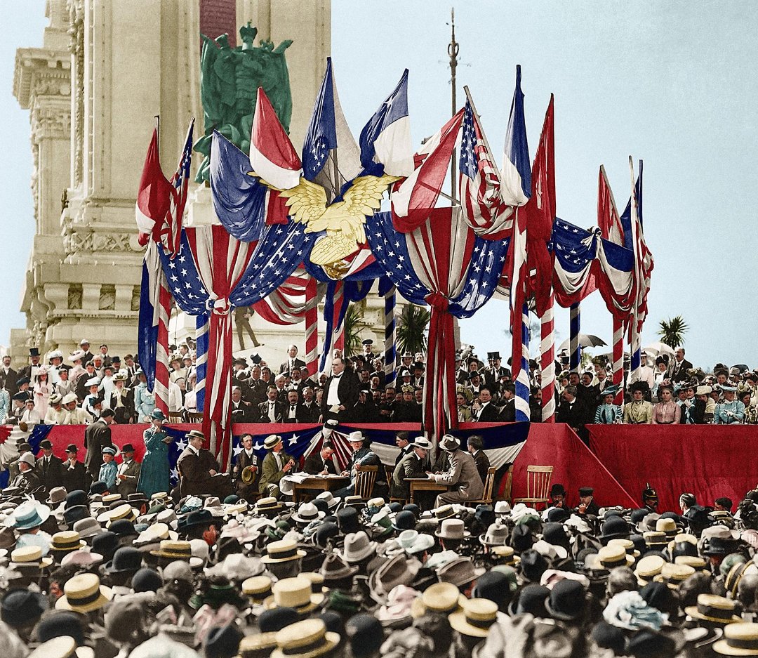 A large crowd gathers at an outdoor event decorated with American flags and bunting. People in early 1900s attire watch speakers on a stage beneath red, white, and blue drapes and an eagle emblem.