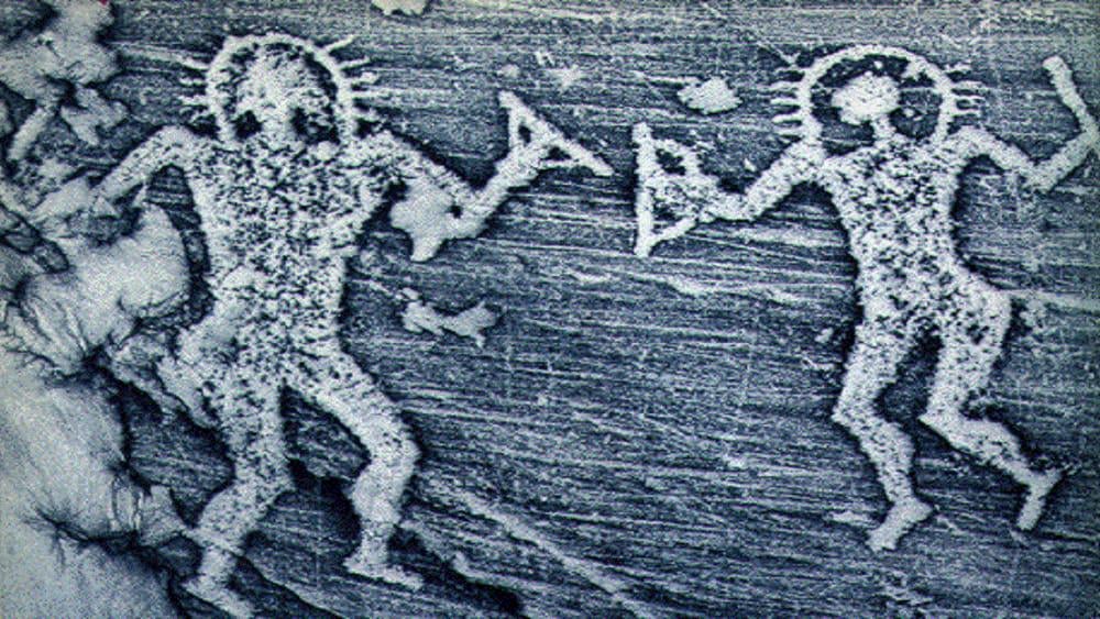 Ancient petroglyph depicting two human-like figures with outstretched arms, holding objects, and surrounded by radiating lines, carved into a rough stone surface.