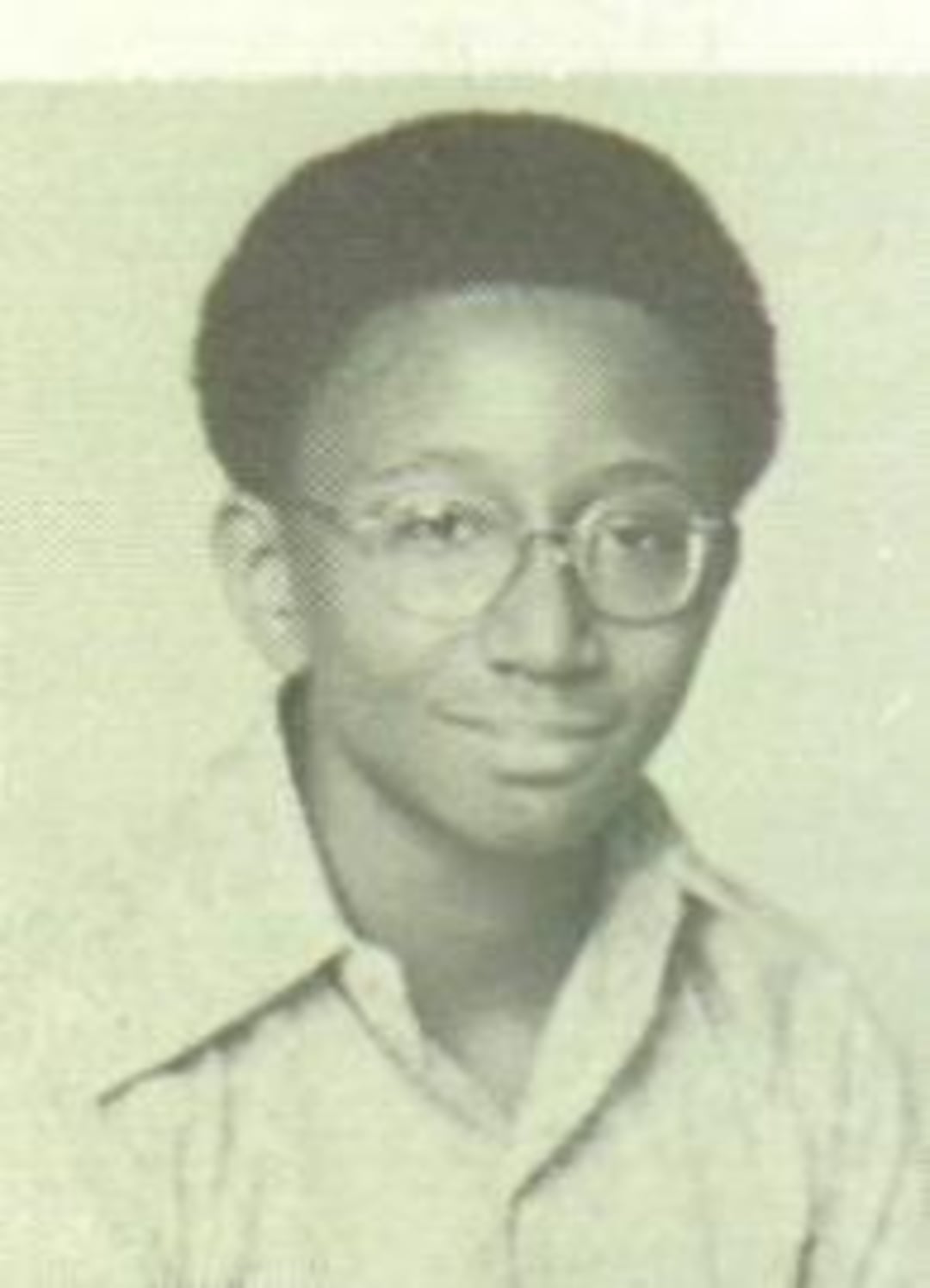 A young person with short curly hair and glasses is wearing a light-colored collared shirt and smiling slightly in this black and white portrait photograph.
