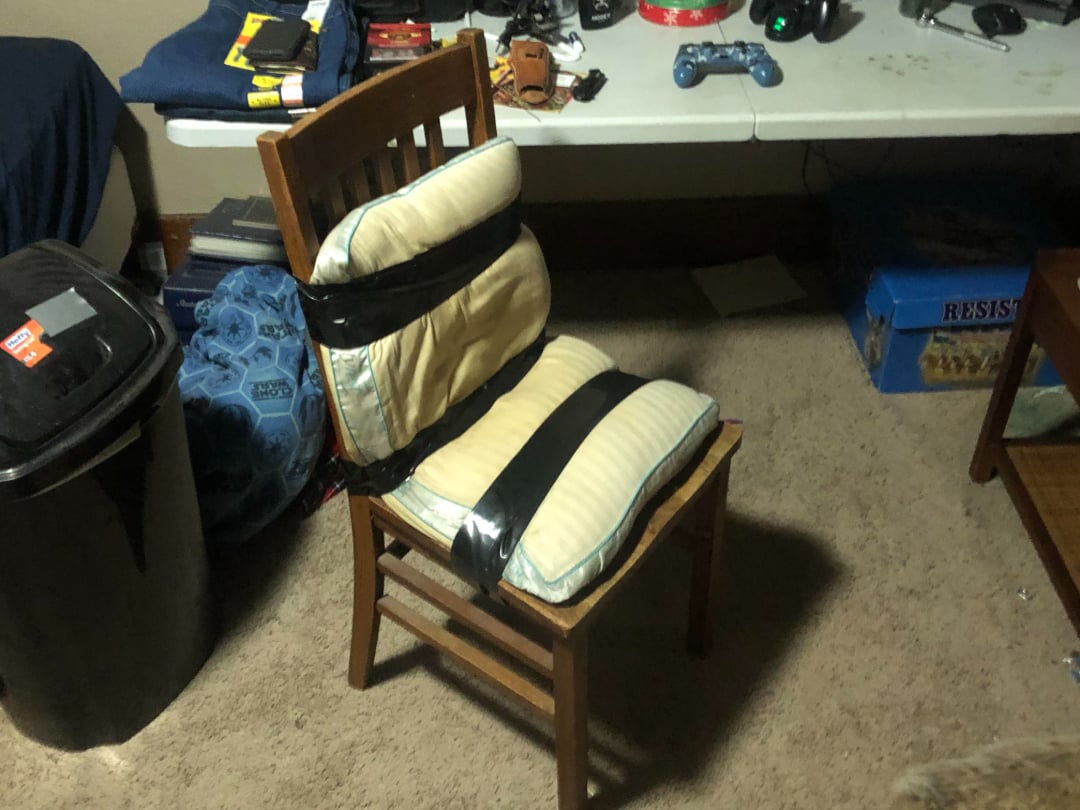 A wooden chair with two bed pillows taped to the seat and backrest using black duct tape, sitting on a carpeted floor near a table with electronics and other items.