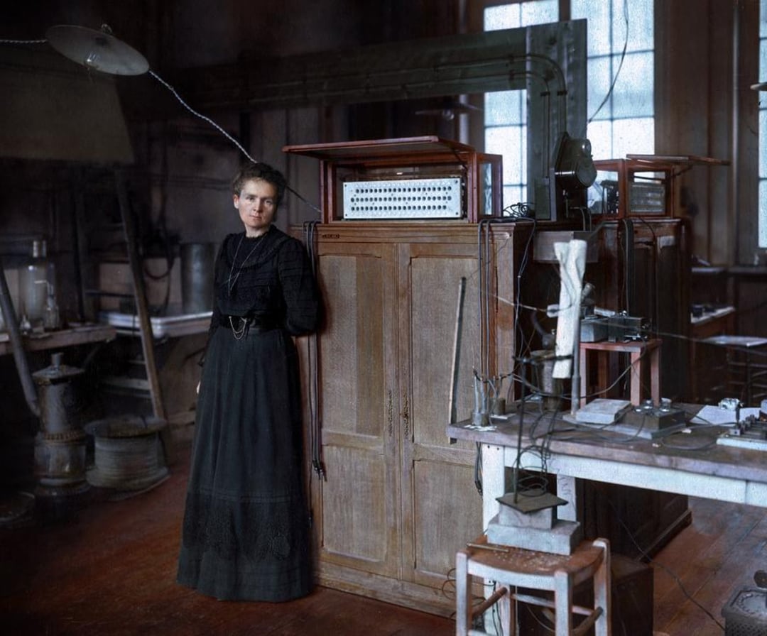 A woman in a long dark dress stands next to scientific equipment and large cabinets in an old laboratory with tall windows and wooden floors. Various instruments and wires are seen on nearby tables.