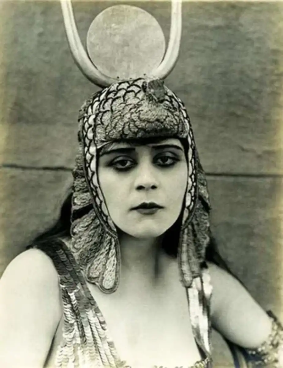 A woman in elaborate ancient Egyptian-style costume and headdress with a large disk and horns, gazes directly at the camera against a plain background in a vintage black-and-white photograph.