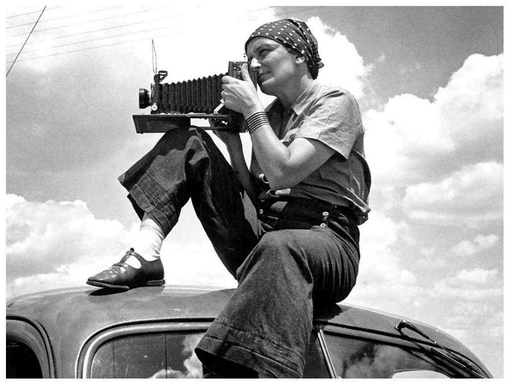 A person wearing a headscarf, cuffed pants, and flat shoes sits on top of a car, using a vintage camera to take a photo, with a partly cloudy sky in the background.
