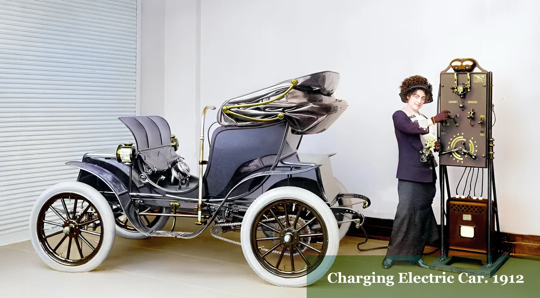 A woman in early 1900s attire stands beside a vintage electric car, plugging it into a large wall-mounted charger. Text on the image reads: "Charging Electric Car: 1912.