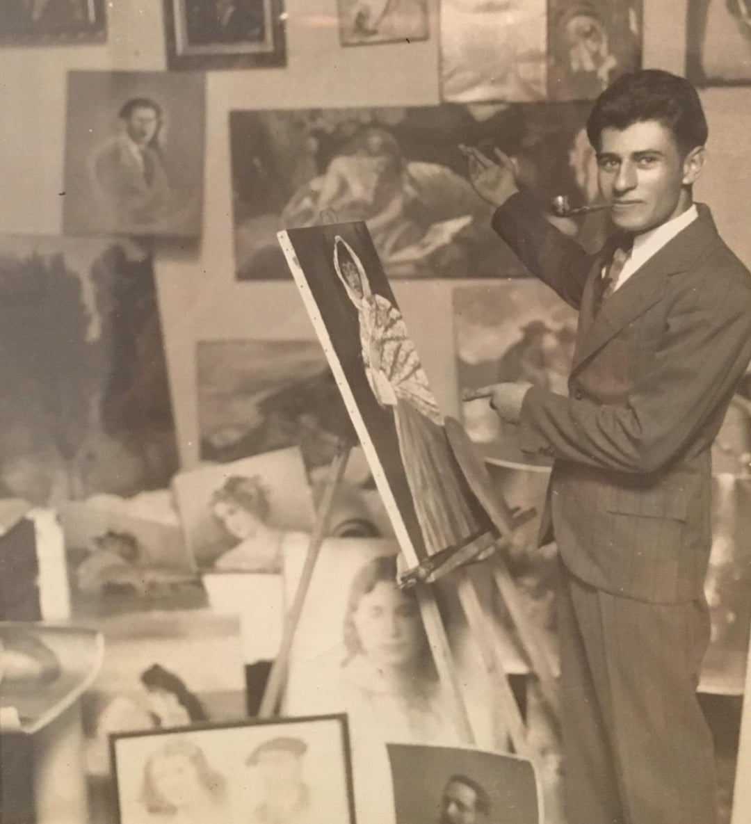 A man in a suit stands beside an easel, painting a portrait. The wall behind him is filled with various portraits and artworks, creating an artistic and creative atmosphere. The photo appears to be vintage or historical.