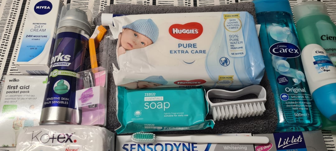 A collection of personal care items, including wipes, deodorant, soap, toothbrushes, toothpaste, day cream, face wipes, sanitary pads, a nail brush, and a small first aid kit, all arranged on a patterned surface.