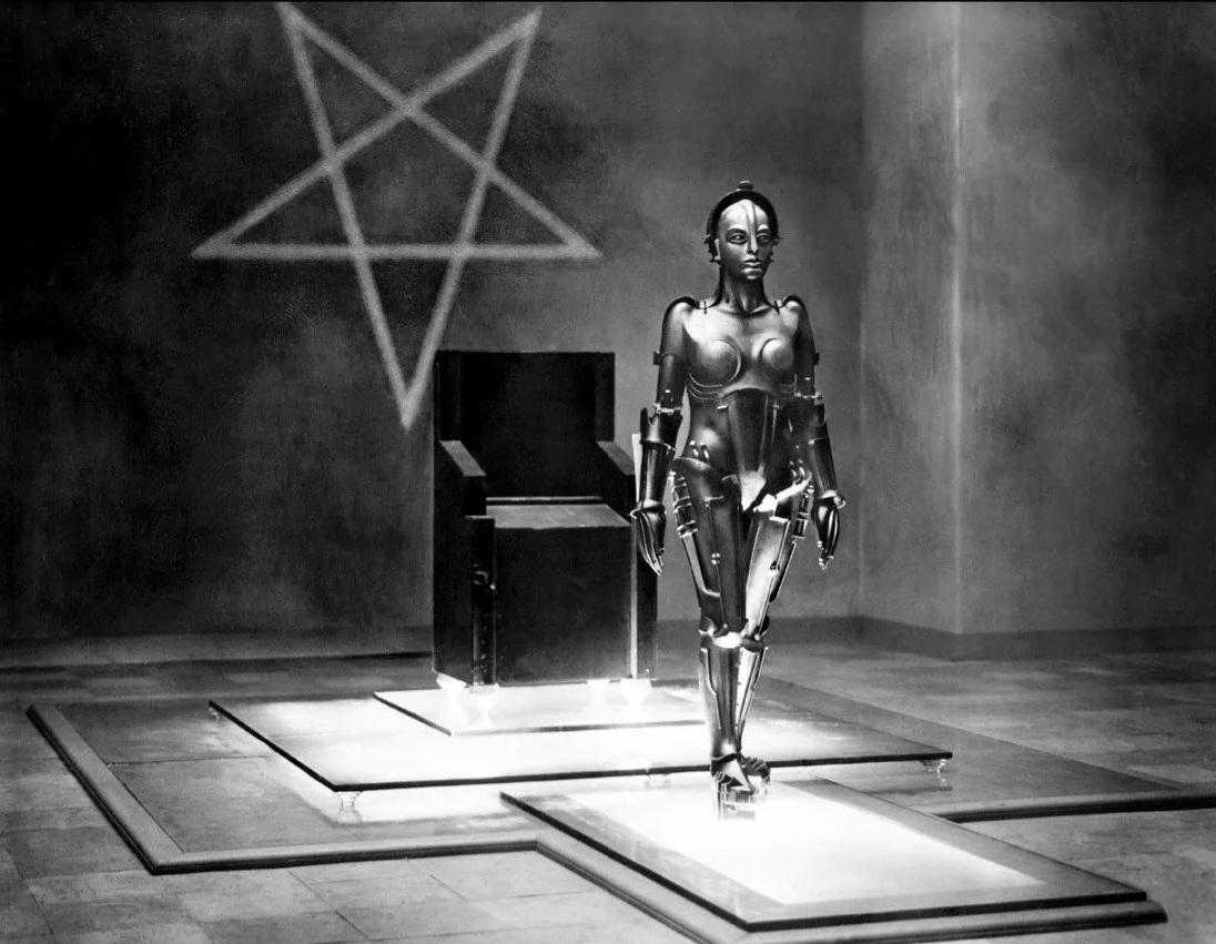 A humanoid robot stands on a lit platform in a stark room with a black empty throne and a large pentagram symbol on the wall behind it. The scene is in black and white.