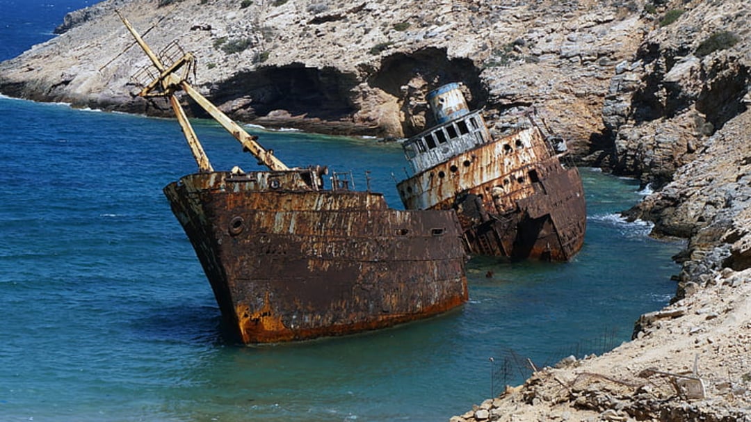 A large, rusted shipwreck rests partially submerged in clear blue water near a rocky coastline, with cliffs and sparse vegetation in the background.