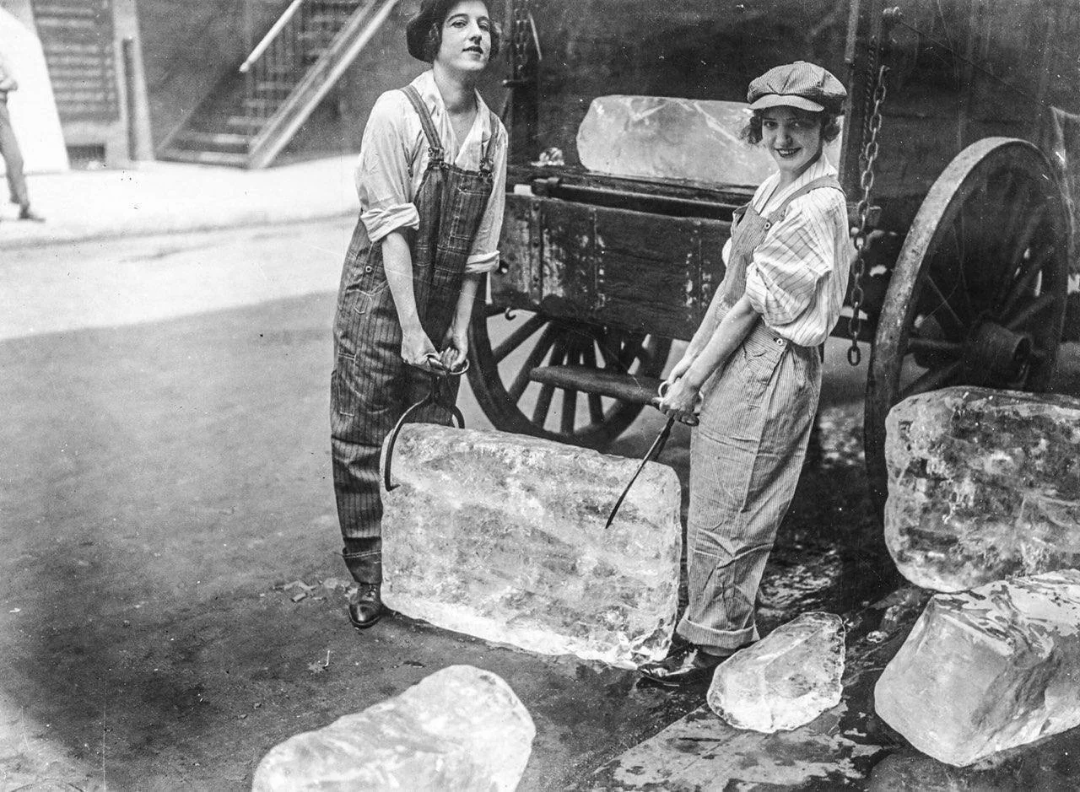 Two women in overalls and caps use ice tongs to carry a large block of ice on a city street, with an old-fashioned wagon and more ice blocks nearby.