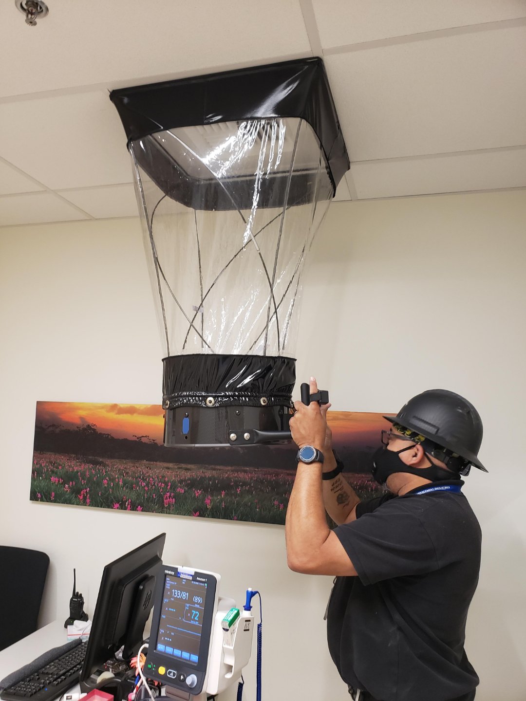 A person wearing a hard hat and mask uses a device to measure airflow from a ceiling vent, with a large plastic airflow capture hood. Medical equipment and a scenic wall photo are visible in the background.