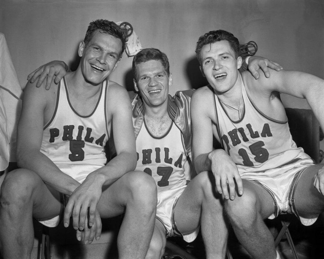Three basketball players in vintage "PHILA" jerseys sit closely together on a bench, smiling and posing for the camera. They wear sleeveless uniforms with numbers 5, 17, and 15 visible.