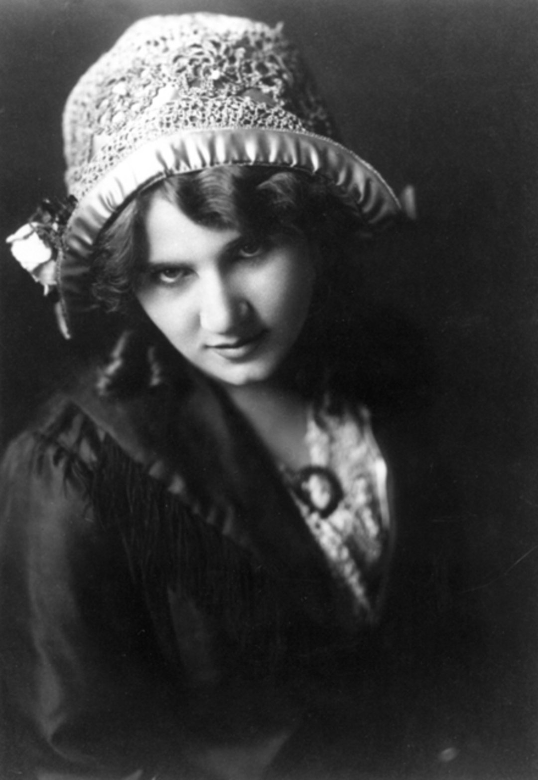 A black-and-white portrait of a woman in vintage attire, wearing a decorative hat with ribbon and lace. She has wavy hair and is looking slightly upward with a subtle, enigmatic expression.