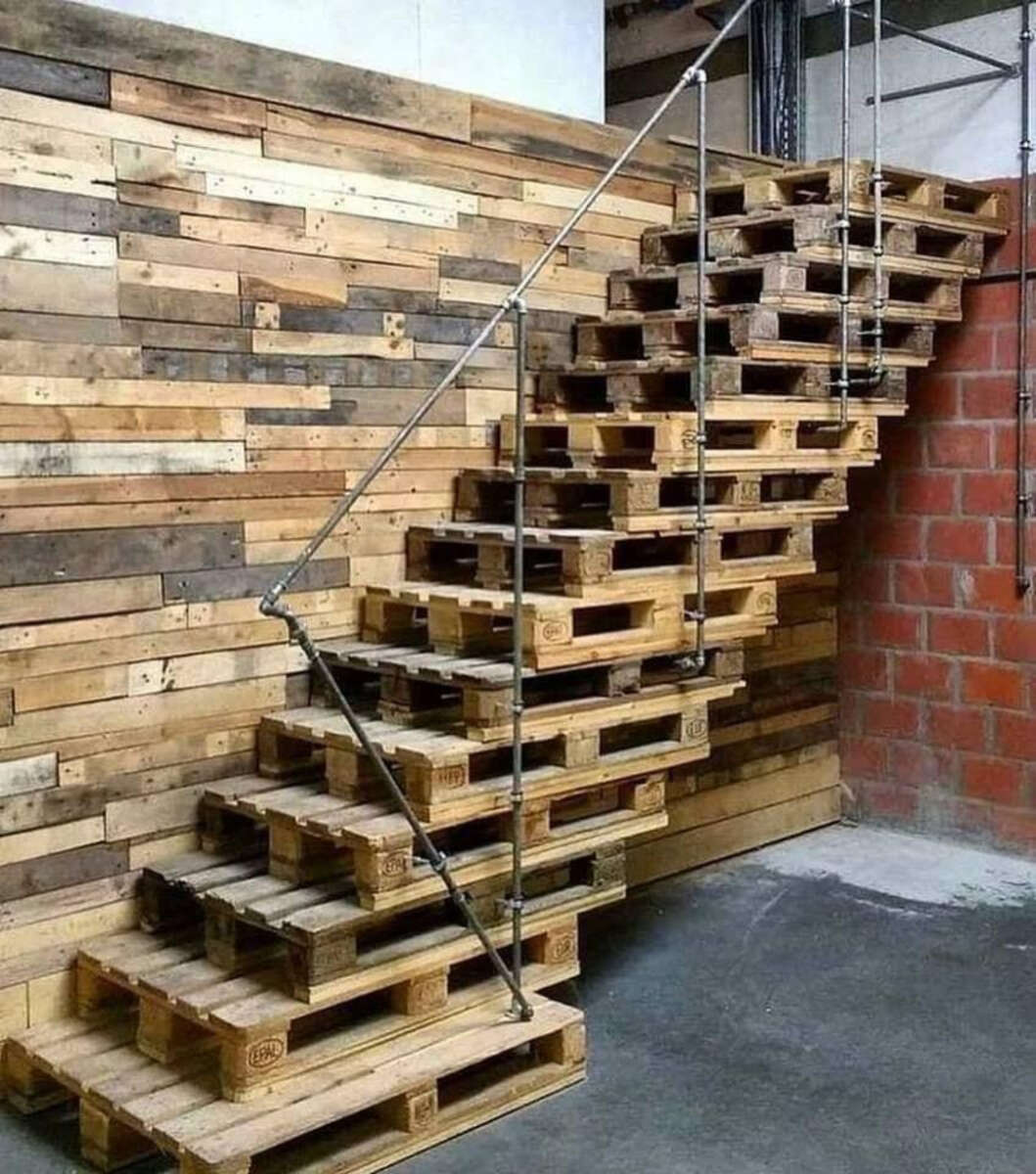 A staircase made from stacked wooden pallets with a metal handrail, set against a wall of horizontal wooden planks and next to a red brick wall on a concrete floor.