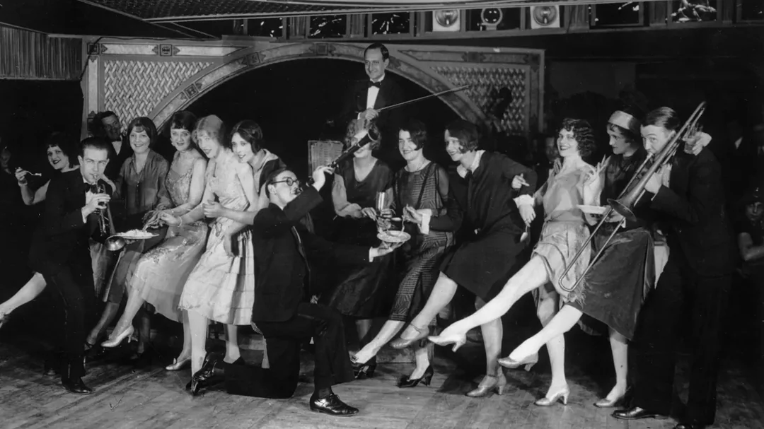 A group of men and women in 1920s attire dance and pose energetically, raising their legs in unison on a dance floor, while a band plays in the background. The atmosphere appears lively and festive.
