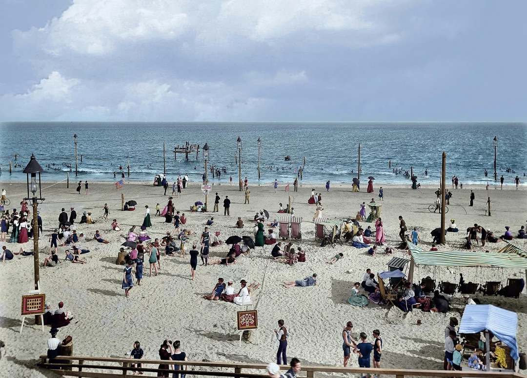 A busy beach scene with many people relaxing on the sand, walking, and playing near the shoreline. Colorful tents and canopies are set up, and the ocean stretches out under a partly cloudy sky.