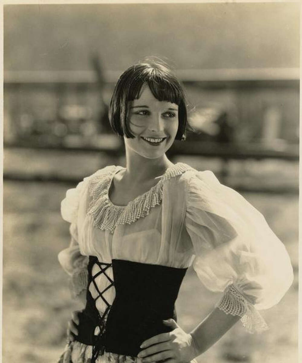 A woman in vintage clothing with a lace-up bodice and puffed sleeves stands outdoors, smiling and looking to the side. The image is in black and white and has an old-fashioned, early 20th-century style.