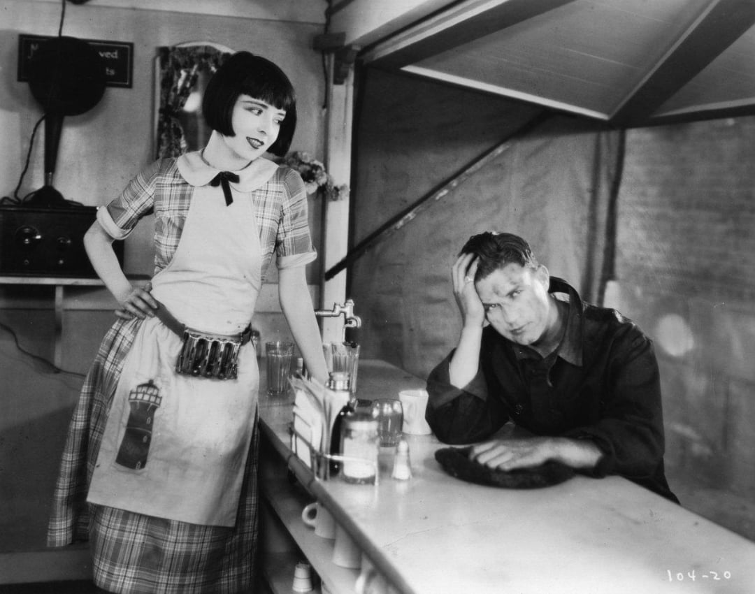 A woman in a plaid dress and apron stands behind a diner counter, looking at a weary man sitting with his head in his hand. Glass bottles and a napkin holder are visible on the counter. The scene appears to be from an old film.