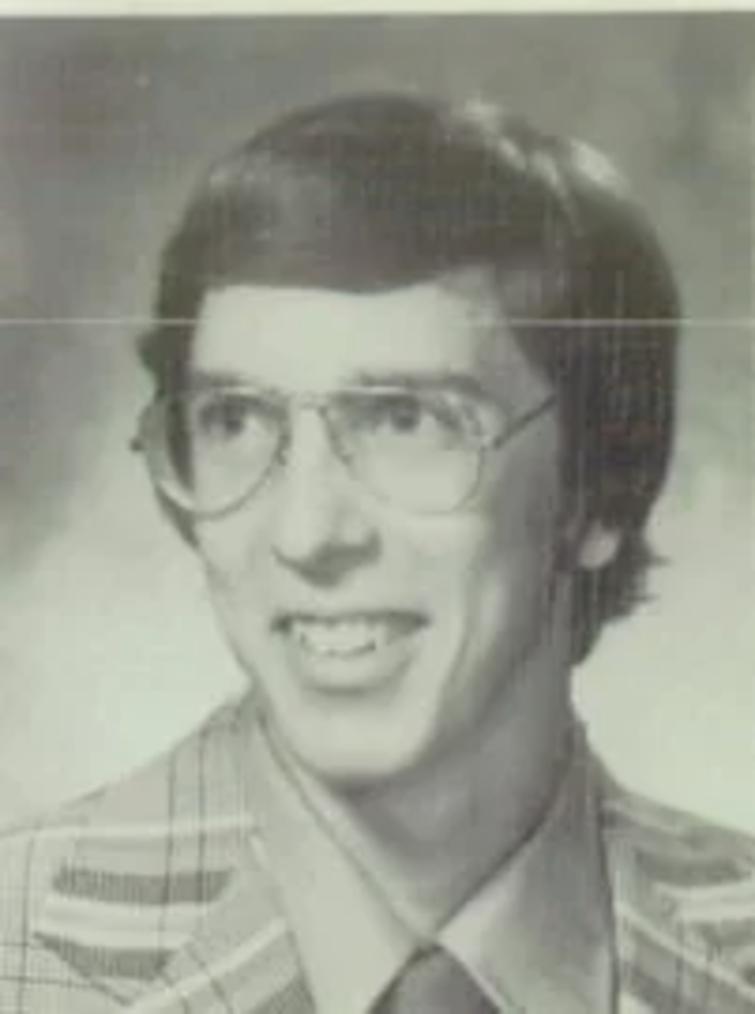 A young man wearing large glasses, a plaid jacket, and a collared shirt is smiling in this black-and-white portrait photograph.