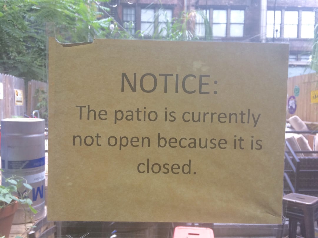 A sign reads, "NOTICE: The patio is currently not open because it is closed." The sign is posted on a window with a view of a fenced outdoor area.