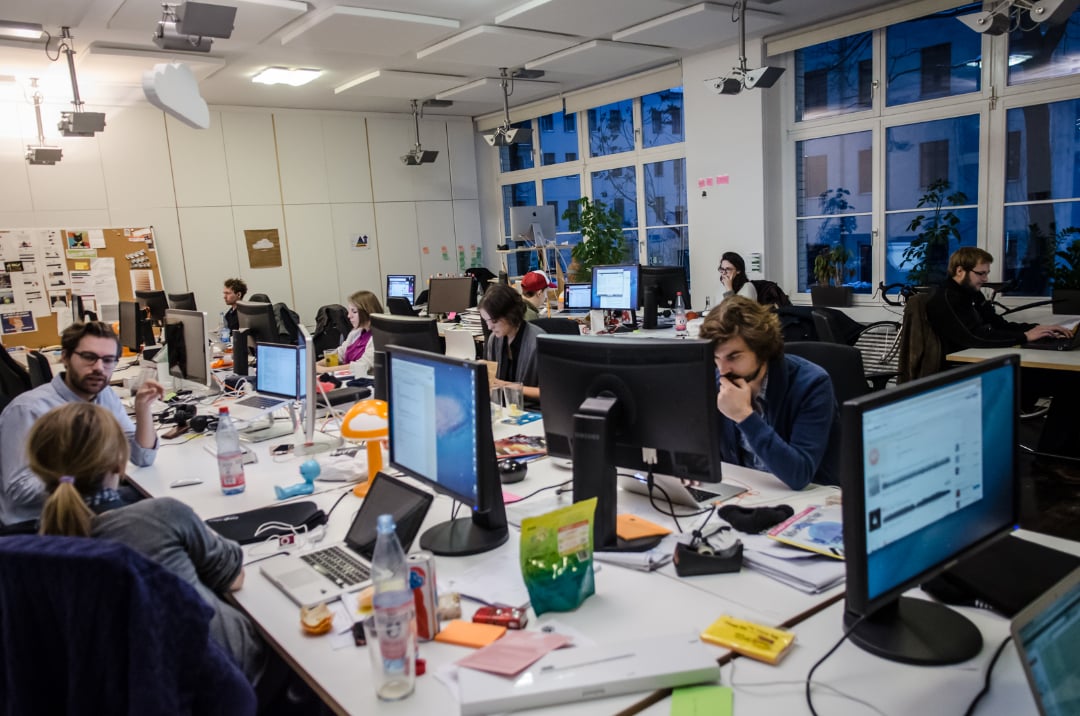 An open-plan office with several people working at desks, using computers. Papers, drinks, and office supplies are scattered on the desks. Large windows line one wall, letting in natural light. The atmosphere appears busy and collaborative.