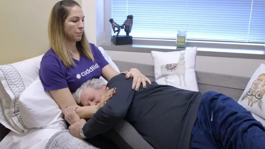 A woman wearing a purple shirt sits on a couch, cradling and comforting an older man lying on her lap in a cozy room with pillows and soft lighting.