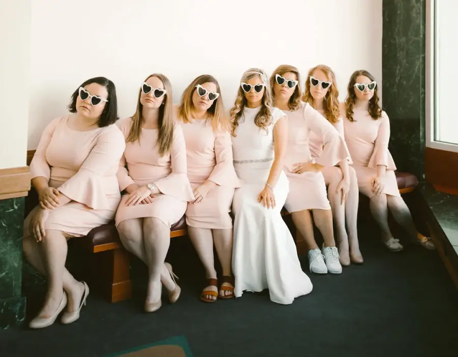Seven women in matching pale pink dresses and heart-shaped sunglasses sit on a bench; one in a white dress suggests she is the bride, while the others are likely bridesmaids. All are posing with relaxed, playful expressions.