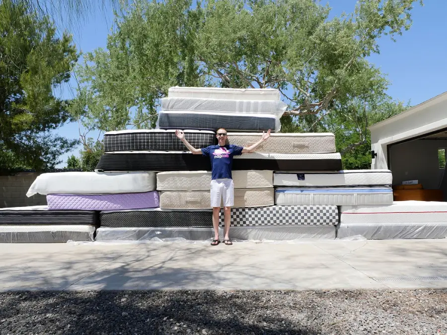 A person stands with arms raised in front of a large stack of mattresses piled outside on a driveway, with trees and a garage visible in the background on a sunny day.