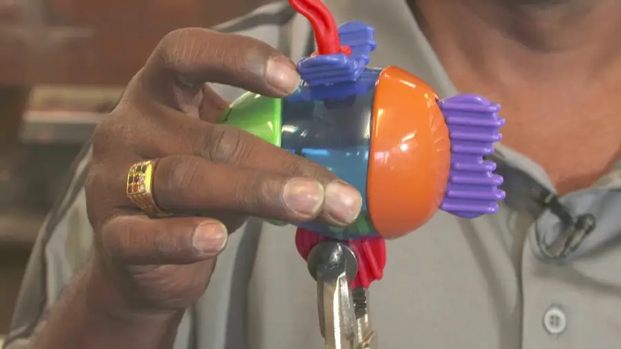 A person holds a colorful plastic toy fish with purple fins and orange, green, and blue body parts, while gripping it with pliers. The person wears a gold ring and a grey shirt.