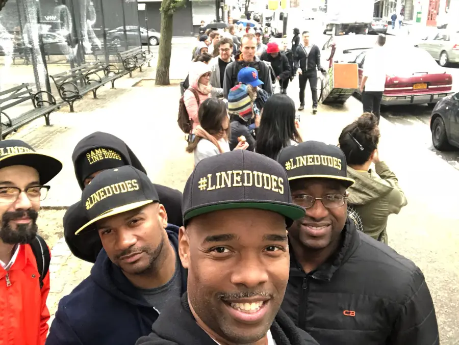 A group of men wearing matching black hats that say "#LINEDUDES" take a selfie while standing in a long line with many people on a city sidewalk.