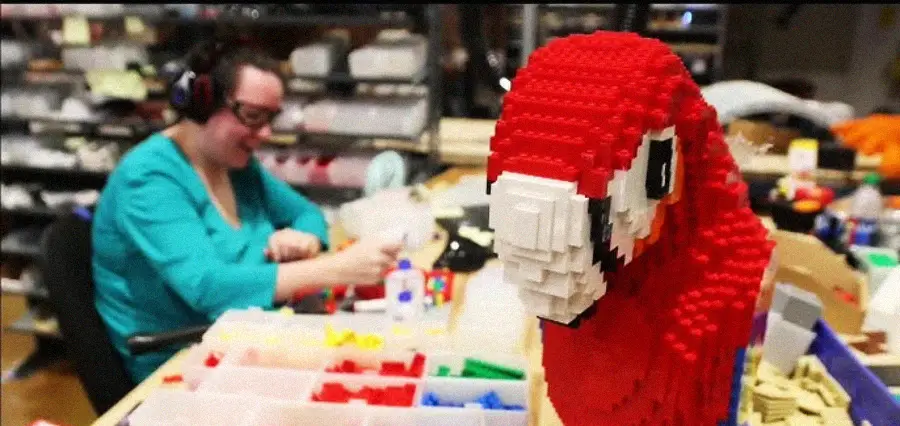 A person sits at a table sorting colorful LEGO bricks while a large red parrot made of LEGO pieces is displayed prominently in the foreground.