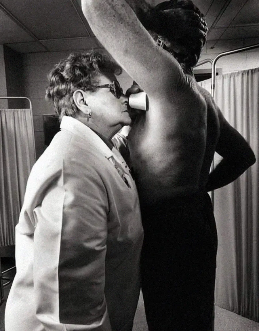 A woman in a white lab coat smells the armpit of a shirtless person raising their arm, possibly conducting an odor test in a clinical setting with privacy curtains in the background.