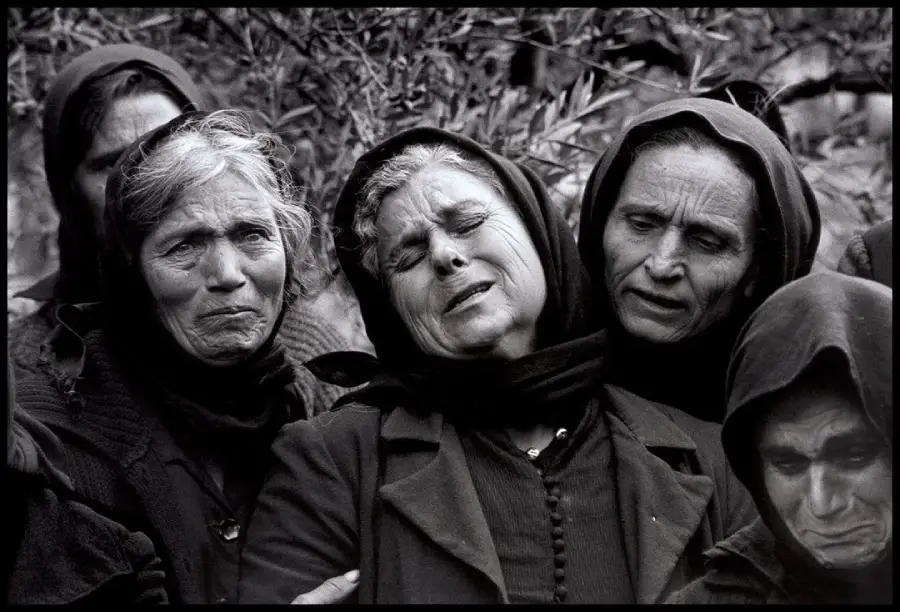 Four women in dark clothing and headscarves stand close together, displaying expressions of deep grief and sorrow. The central woman has her eyes closed, her face contorted in anguish, as others support her.