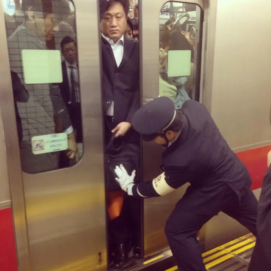 A train station worker pushes a man into a crowded subway car while the train doors attempt to close, with other passengers visible inside.