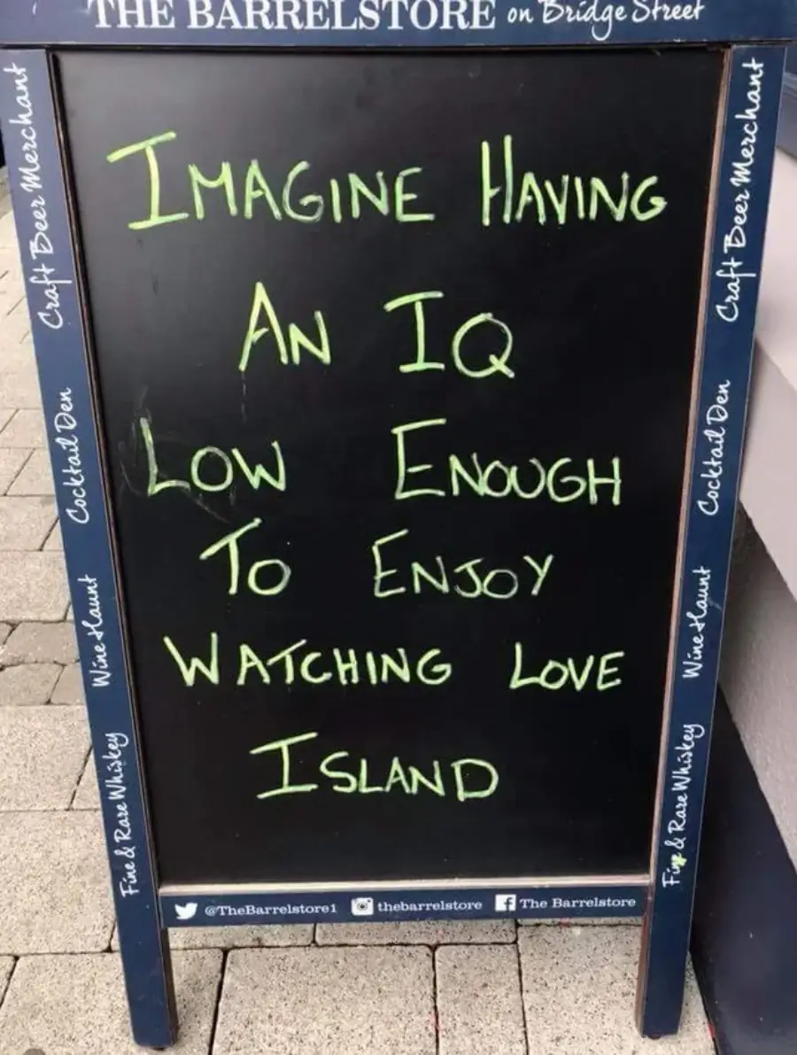 A chalkboard sign reads: "Imagine having an IQ low enough to enjoy watching Love Island," written in green chalk with varied text sizes. The sign is outside The Barrelstore bar.
