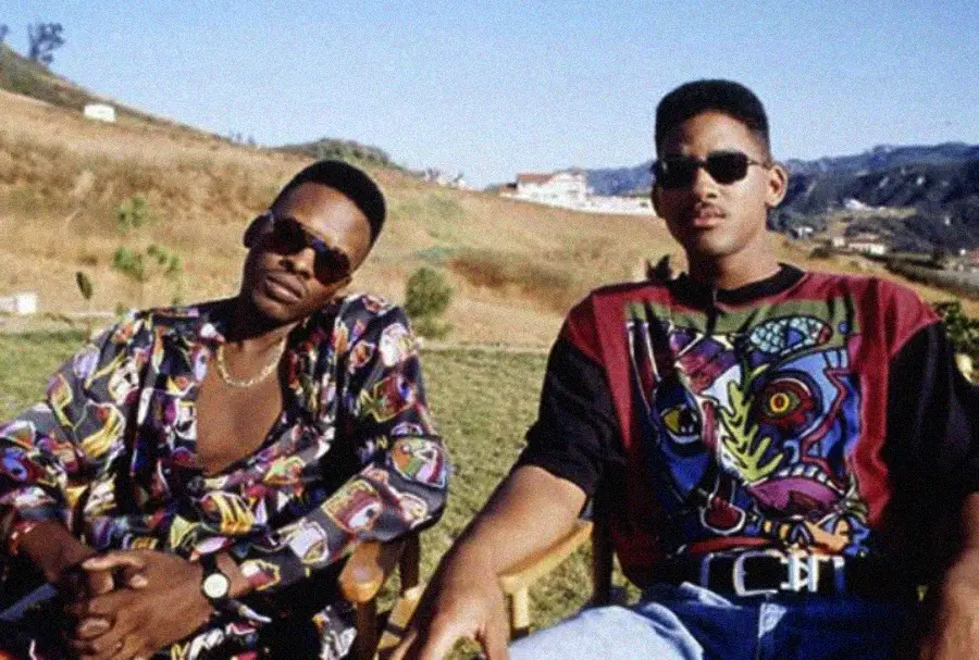 Two men wearing colorful shirts and sunglasses sit outdoors on wooden chairs, with hills and a few houses in the background under a clear sky.