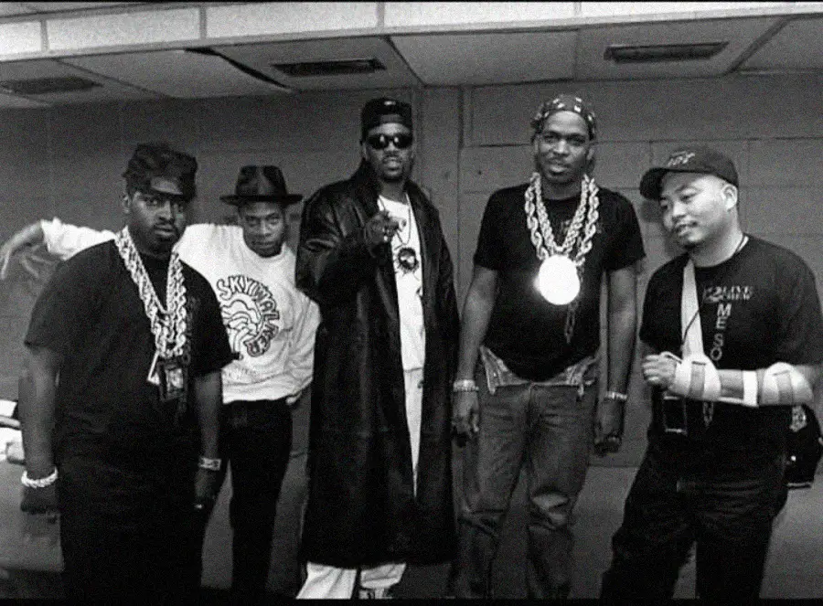 A group of five men stand together indoors, wearing large chain necklaces, hats, and casual clothing; one man wears a long leather coat and sunglasses, while another has his arm in a cast. The scene has a vintage, black-and-white look.