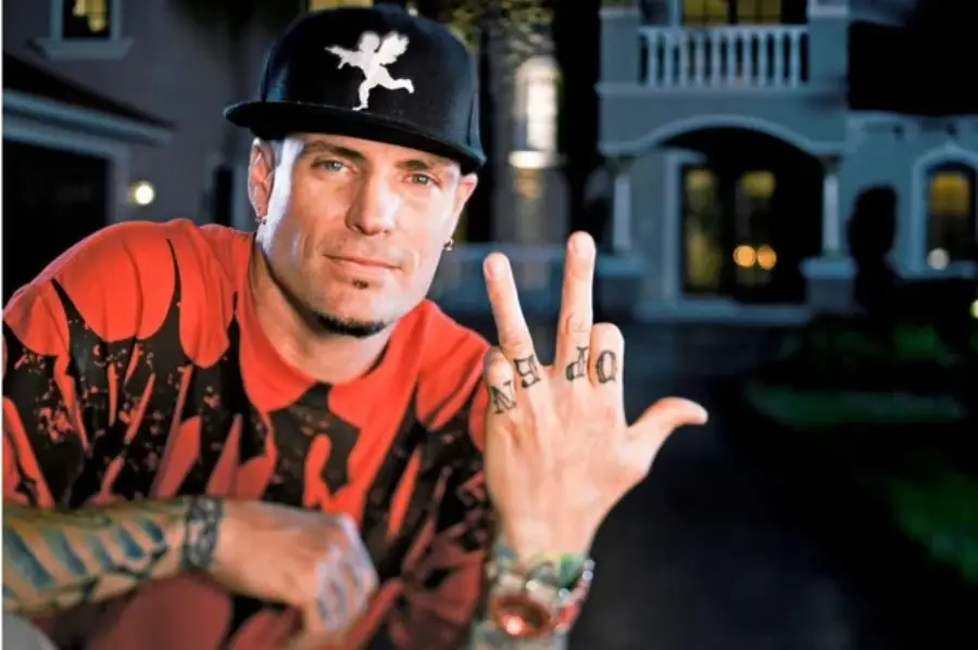 A man wearing a black cap with an angel graphic, a red and black shirt, and showing tattooed fingers that spell "WORD" poses in front of a large, well-lit house at night.