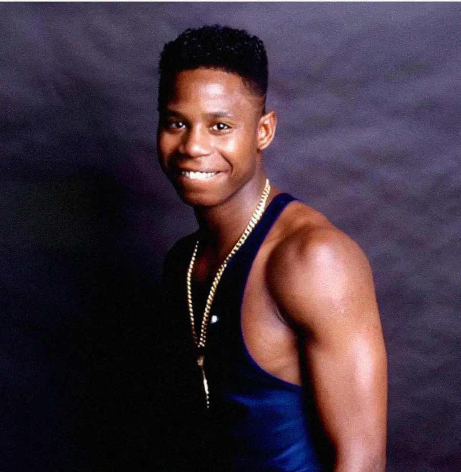 A young man with short hair, wearing a dark tank top and a gold chain necklace, stands and smiles at the camera against a dark, plain backdrop.