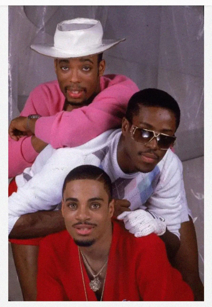 Three men pose in a playful, stacked formation against a light background. The man on top wears a white hat and pink sweater, the middle man wears sunglasses and a white glove, and the bottom man wears a red shirt and gold necklace.