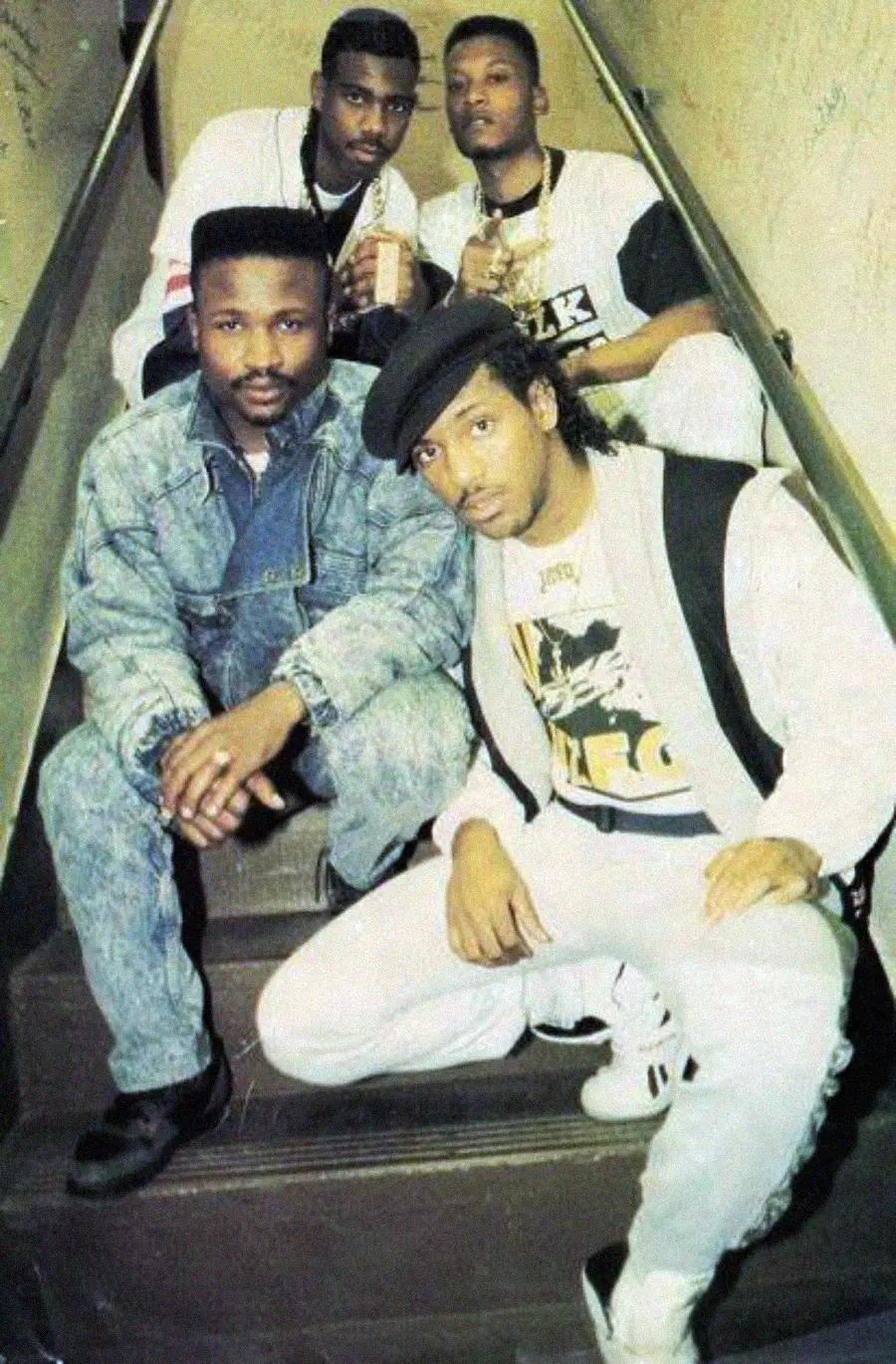 Four men sit on a staircase posing for a photo. They wear late 1980s or early 1990s casual streetwear, including denim, graphic t-shirts, and a cap. The wall behind them is beige with handwritten notes or signatures.