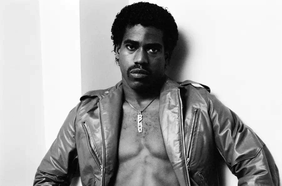 A man with short curly hair and a mustache leans against a wall, wearing an unzipped leather jacket with no shirt underneath. He has a serious expression and a necklace with large letter charms. The photo is in black and white.
